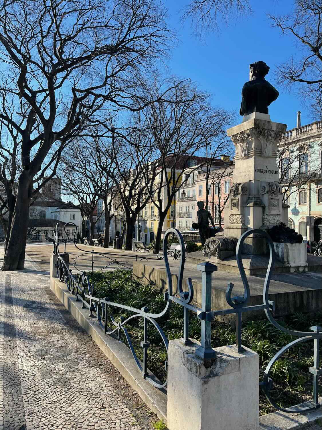 A serene Lisbon neighborhood in the daytime with bare trees, iron-wrought fences, and a statue, showcasing the calm residential side of the city.