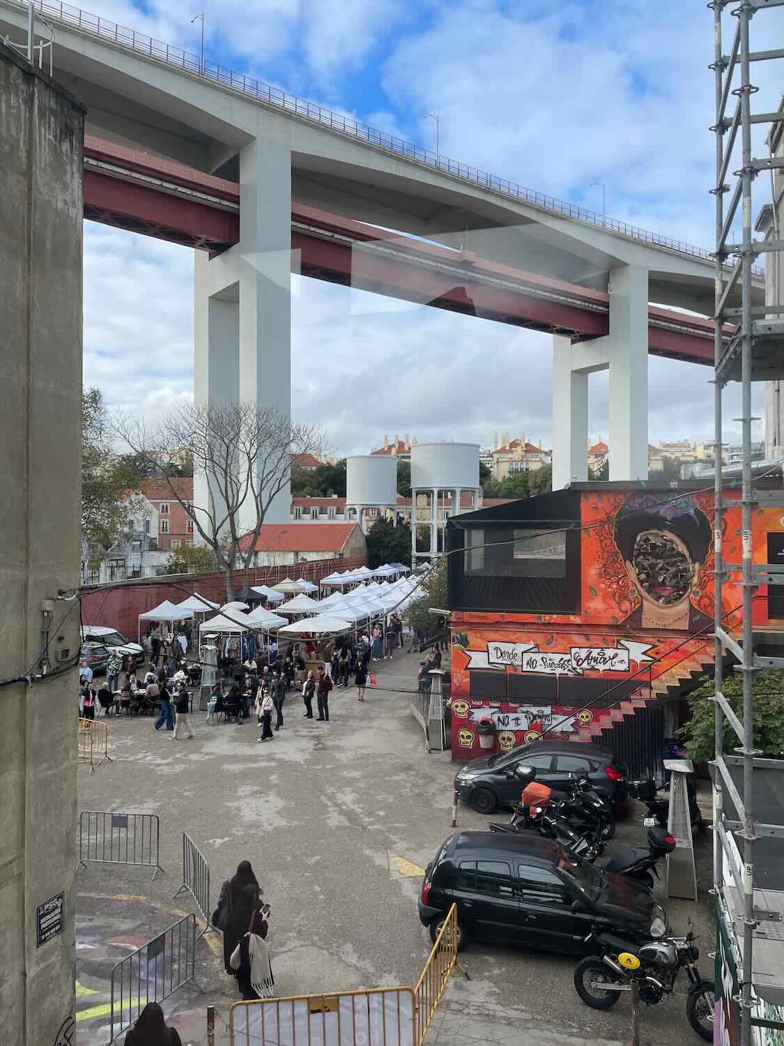 A bustling LX Factory market under the shadow of the overpass in Lisbon, with people exploring stalls and the urban art space, reflecting the city's contemporary culture.