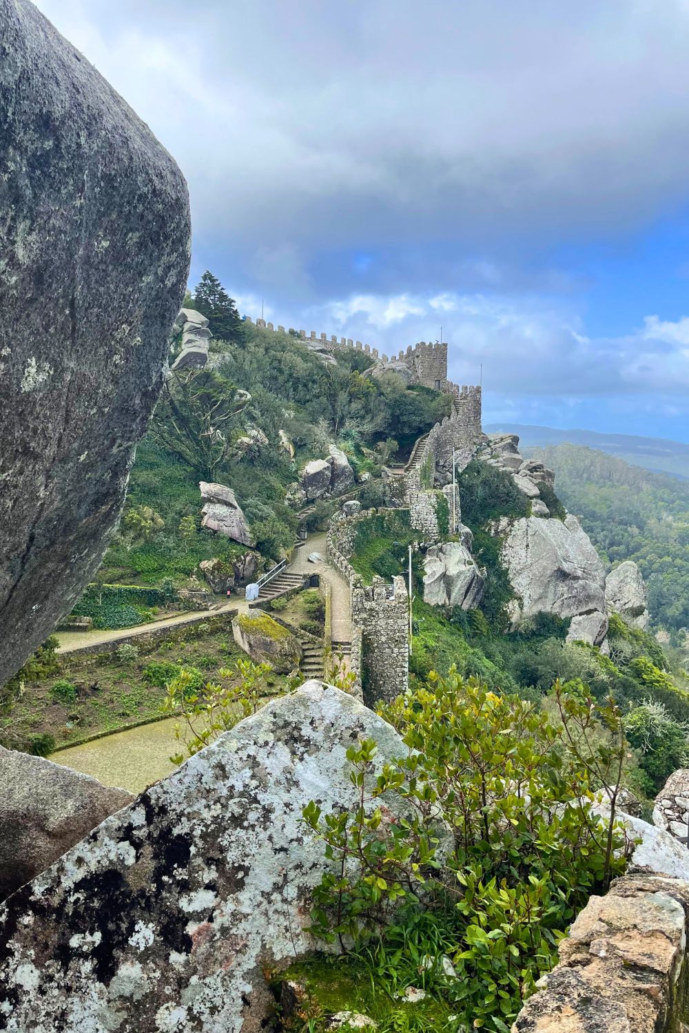 A view of the ancient stone walls and winding staircases of the Moors Castle in Sintra, Portugal, nestled among large boulders and green vegetation, with a hazy sky above and a distant view of the countryside.