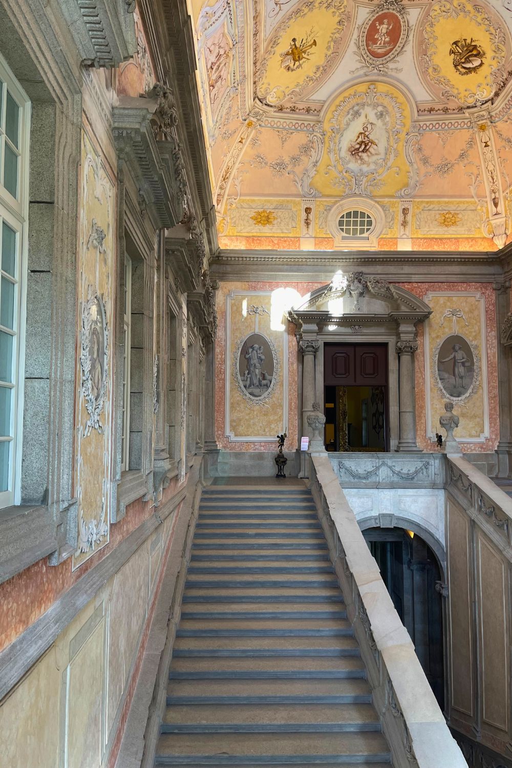 Grand staircase in a historic building in Porto with intricate stucco work, leading to an ornately decorated ceiling with golden motifs and pastel frescoes.