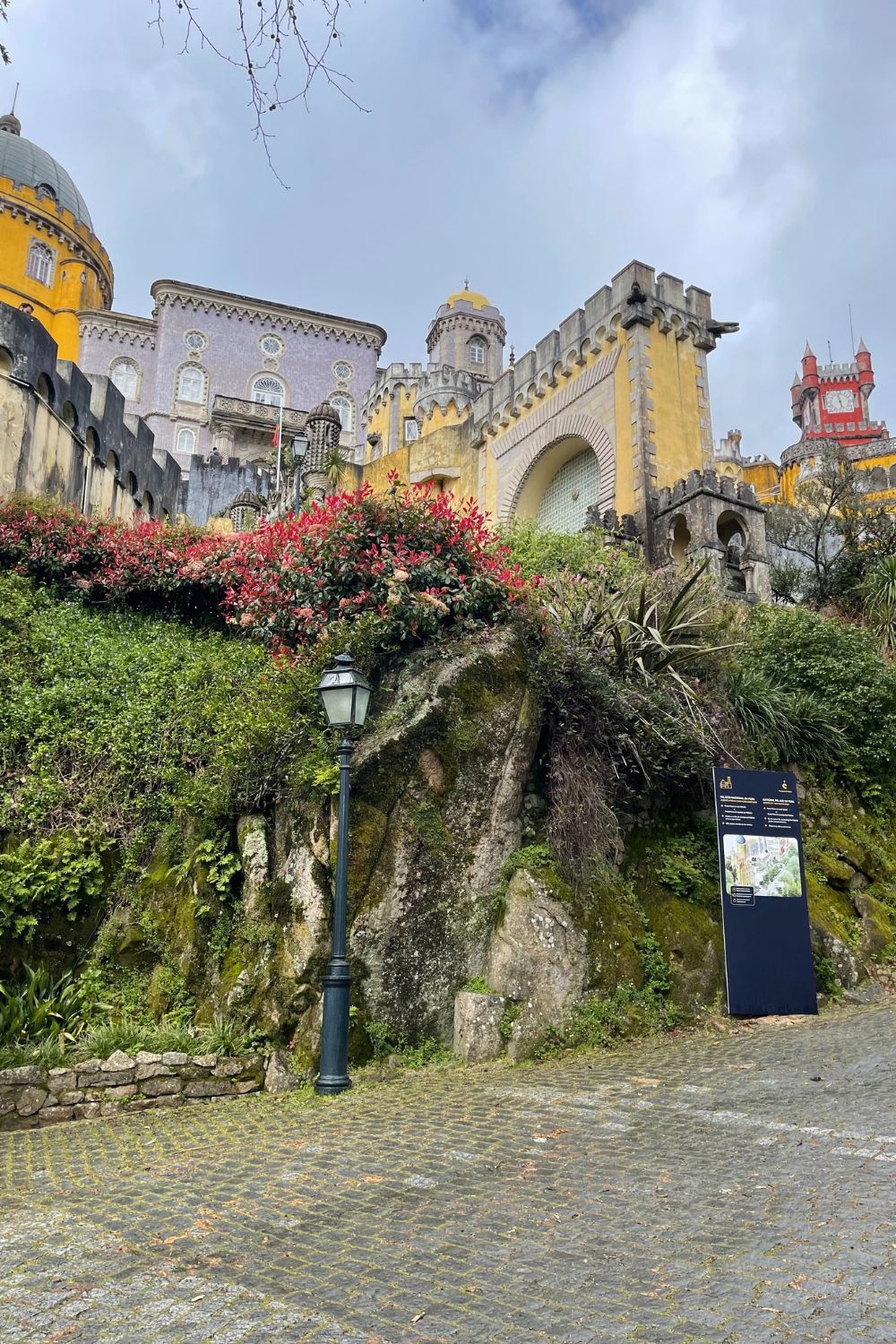 A colorful facade of the Pena Palace in Sintra, Portugal, featuring vibrant yellow and purple walls adorned with traditional Portuguese tiles, surrounded by lush greenery and a vintage street lamp in the foreground.