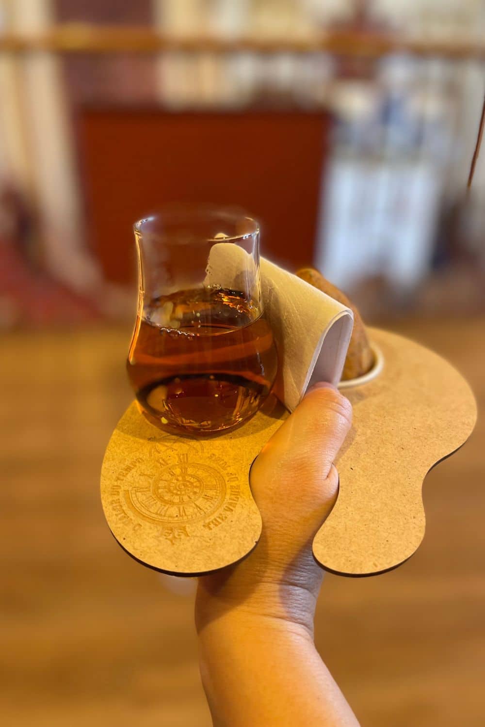 A hand holding a glass of amber-colored port wine with a coaster, set against a blurred background of a wine cellar's interior.