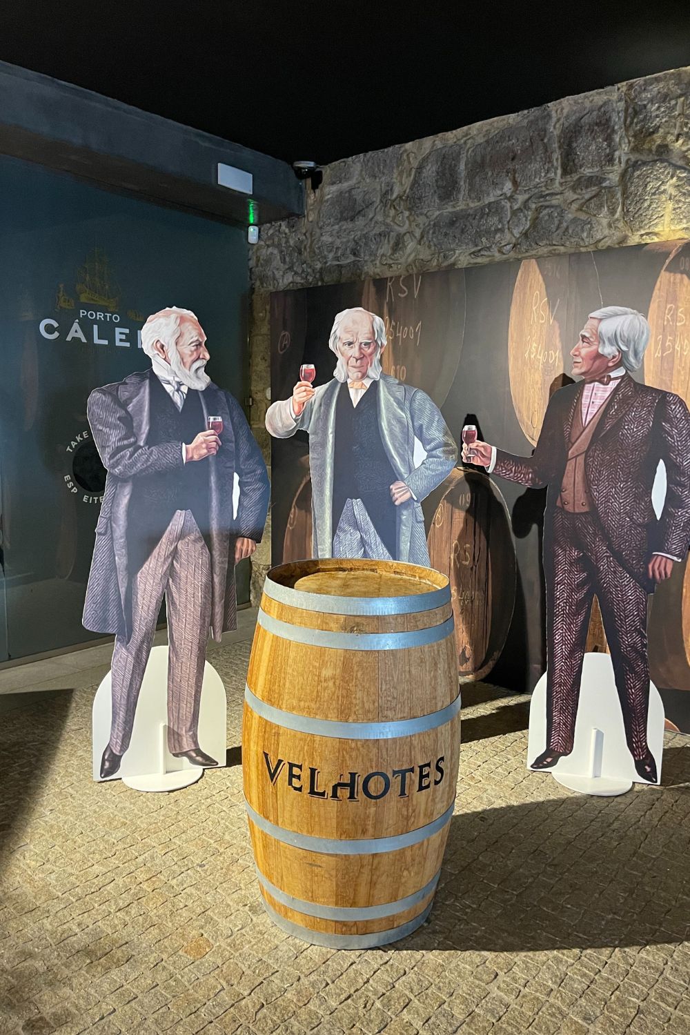 Life-size cardboard cutouts of historical figures holding wine glasses, standing next to a wooden port wine barrel, inside a winery exhibition.
