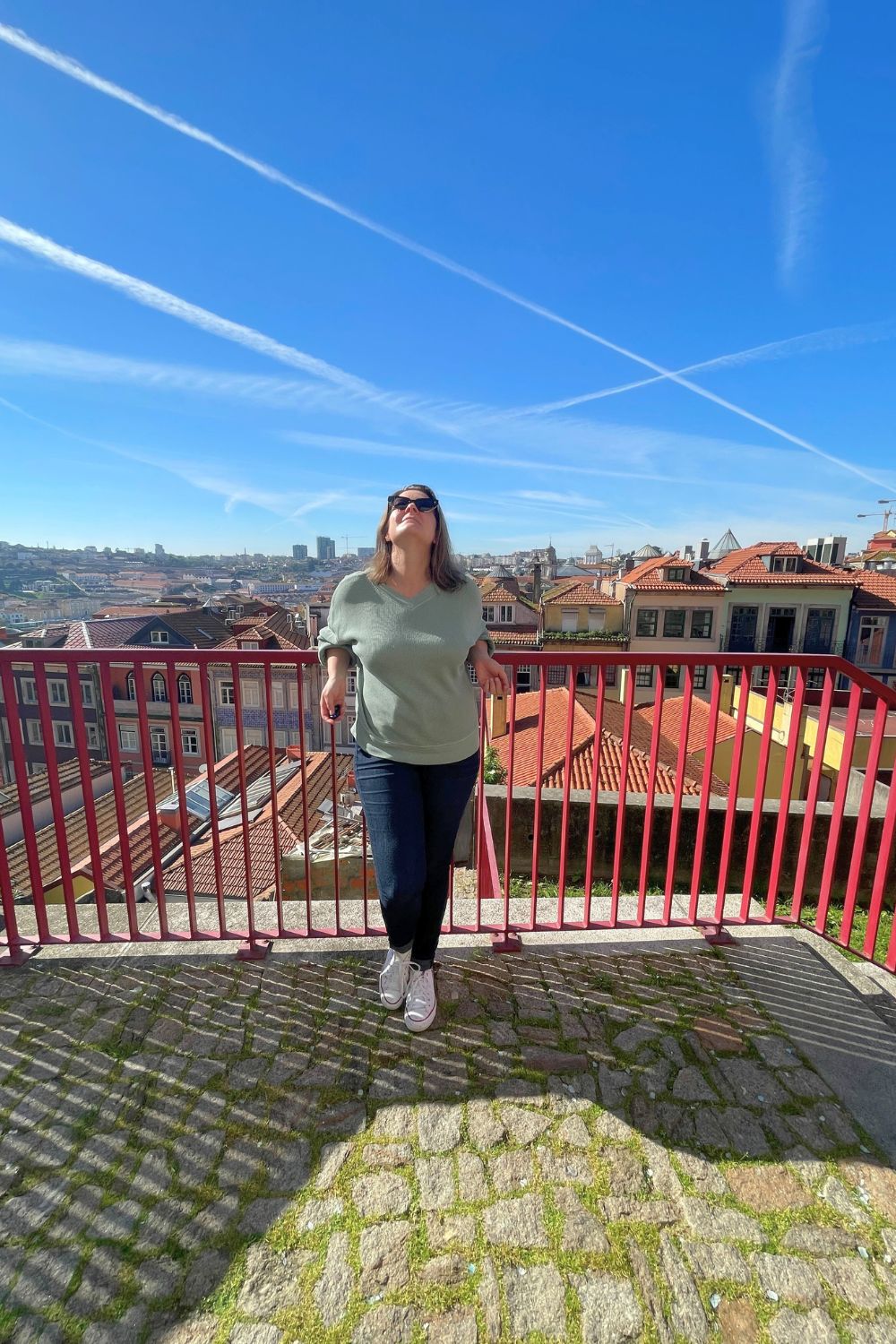 A woman standing on a viewpoint, smiling as she enjoys the expansive view of the red-roofed cityscape of Porto under a bright blue sky with airplane contrails
