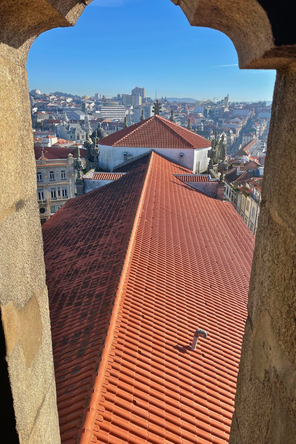 View from a stone archway over the terracotta rooftops of Porto, with the city's buildings stretching into the distance under a clear blue sky.