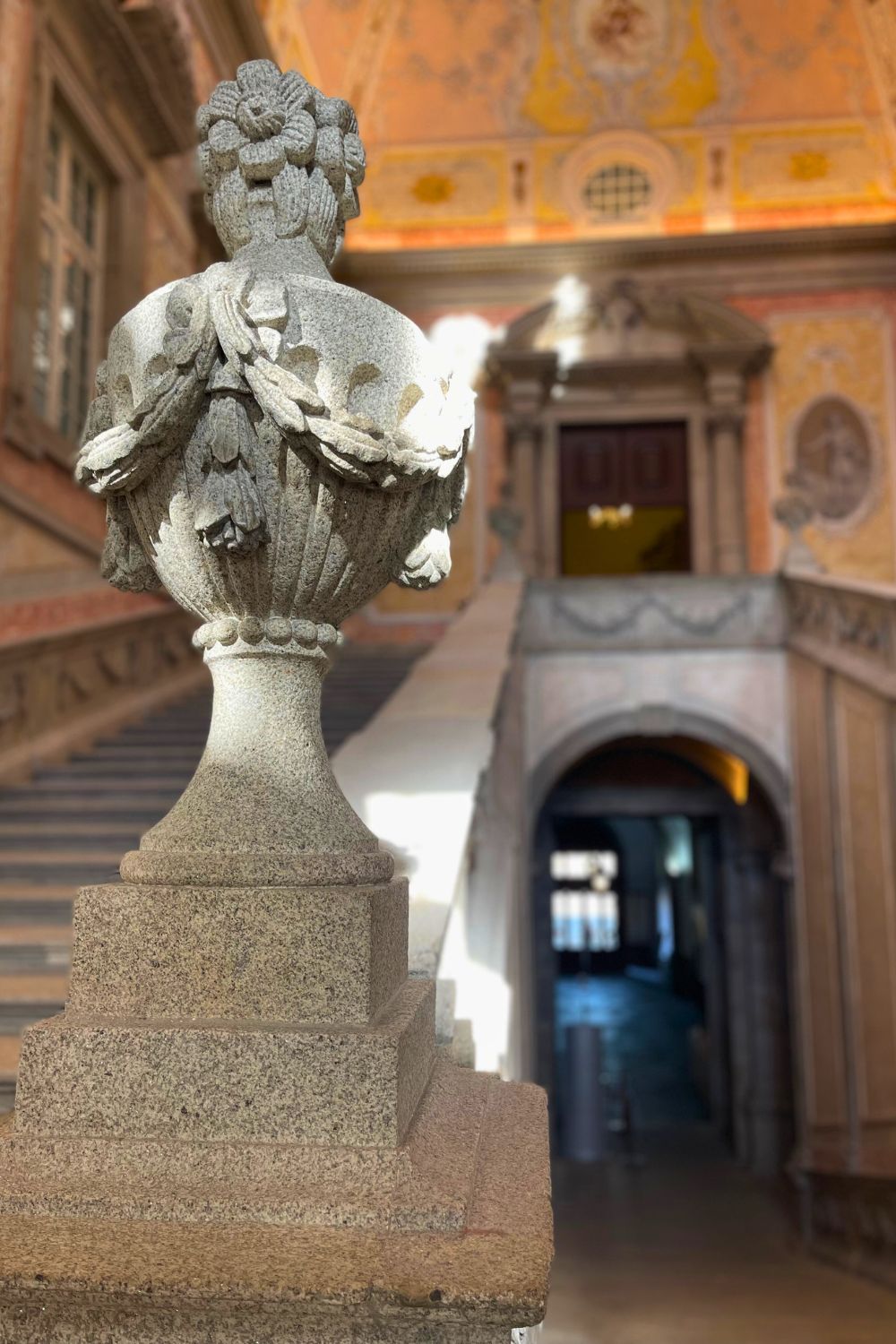Close-up of a decorative stone vase on the balustrade of a grand staircase, with a softly focused background of a richly adorned hallway with golden detailing.