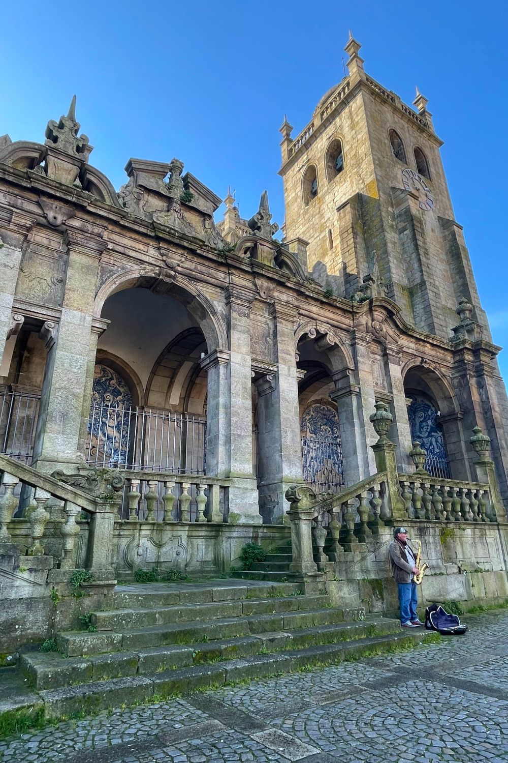 A saxophonist plays in front of the aged stone façade of an ornate church in Porto, under a clear sky, with a backdrop of historic architecture.