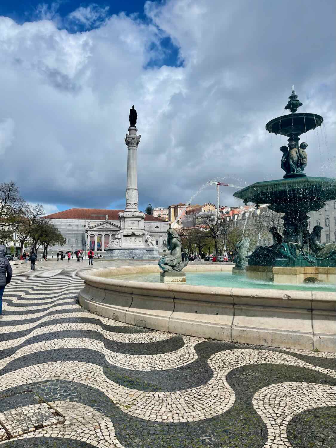Rossio Square in Lisbon, Portugal, on a cloudy day, showcasing the patterned cobblestone pavement, Baroque fountain, and the Pedro IV monument.
