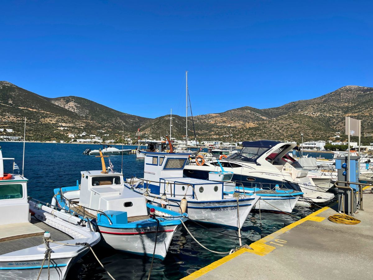 A lively marina in Sifnos with various boats moored along the dock, set against a backdrop of a mountainous landscape and clear blue skies, reflecting the island's nautical charm.