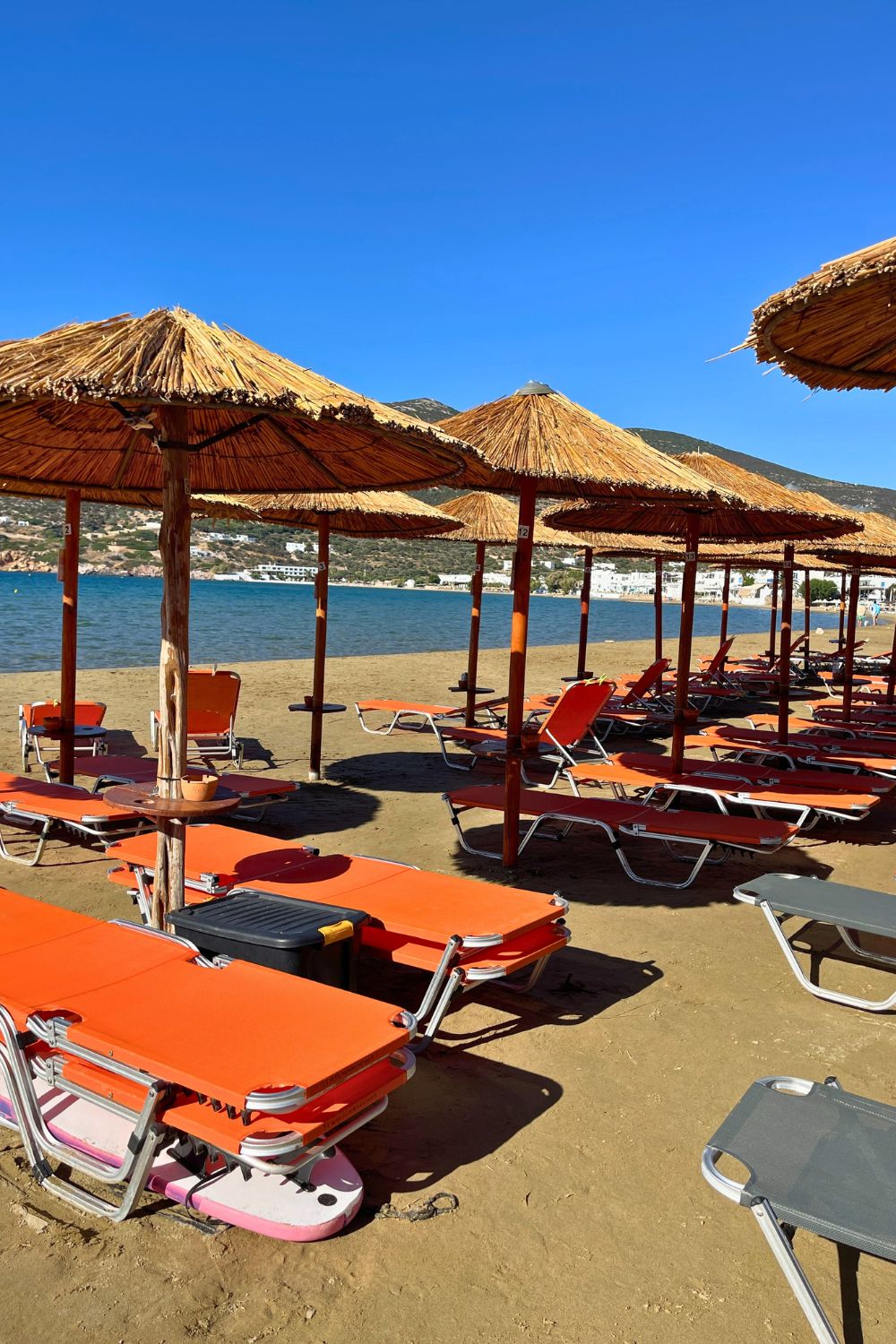 Bright orange beach loungers lined up under thatched parasols on a sandy beach in Sifnos, with the calm Aegean Sea in the background.