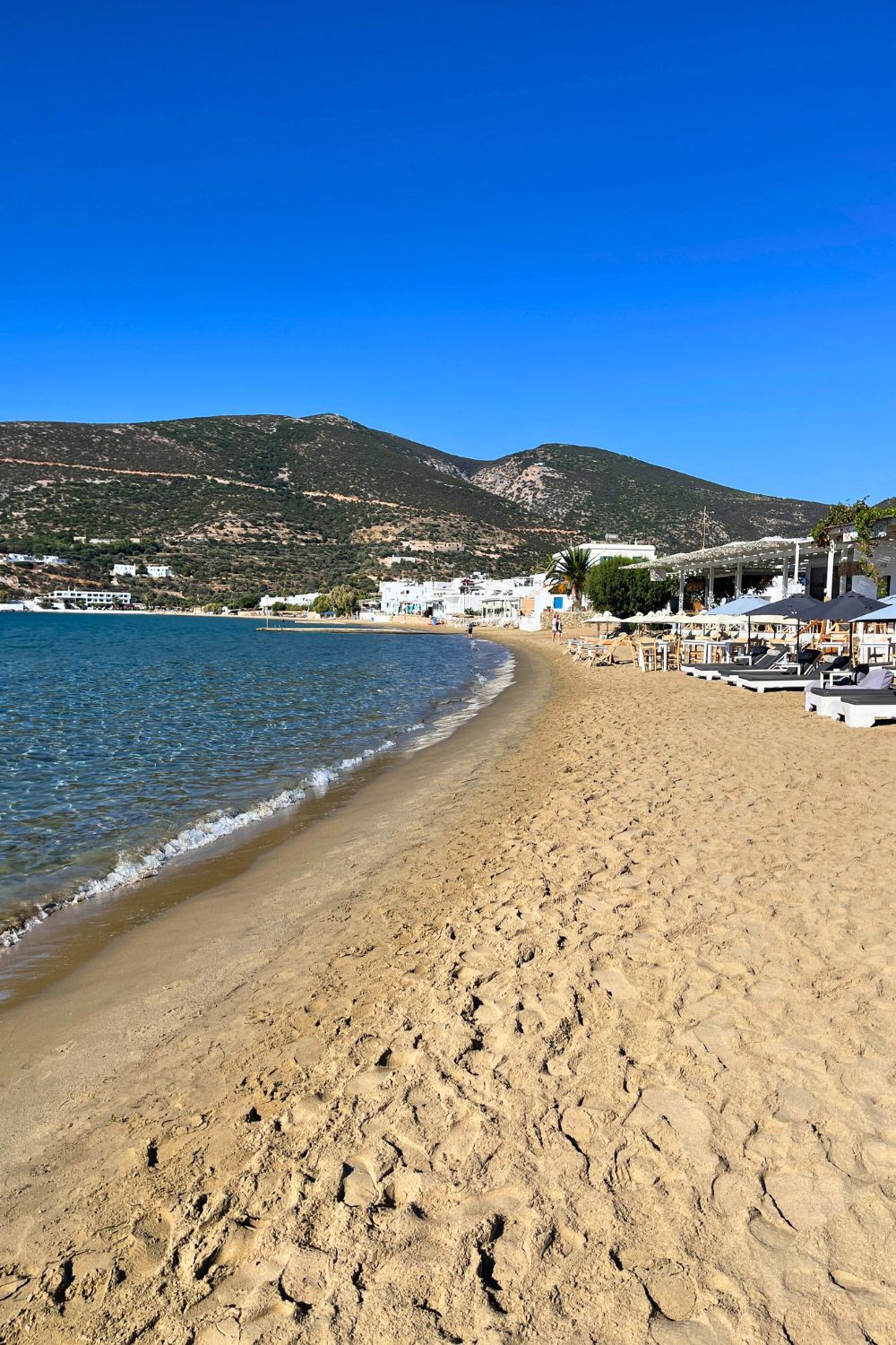 The gentle curve of a sandy beach in Sifnos, lined with beach chairs and umbrellas, set against the backdrop of a tranquil bay and rolling hills.