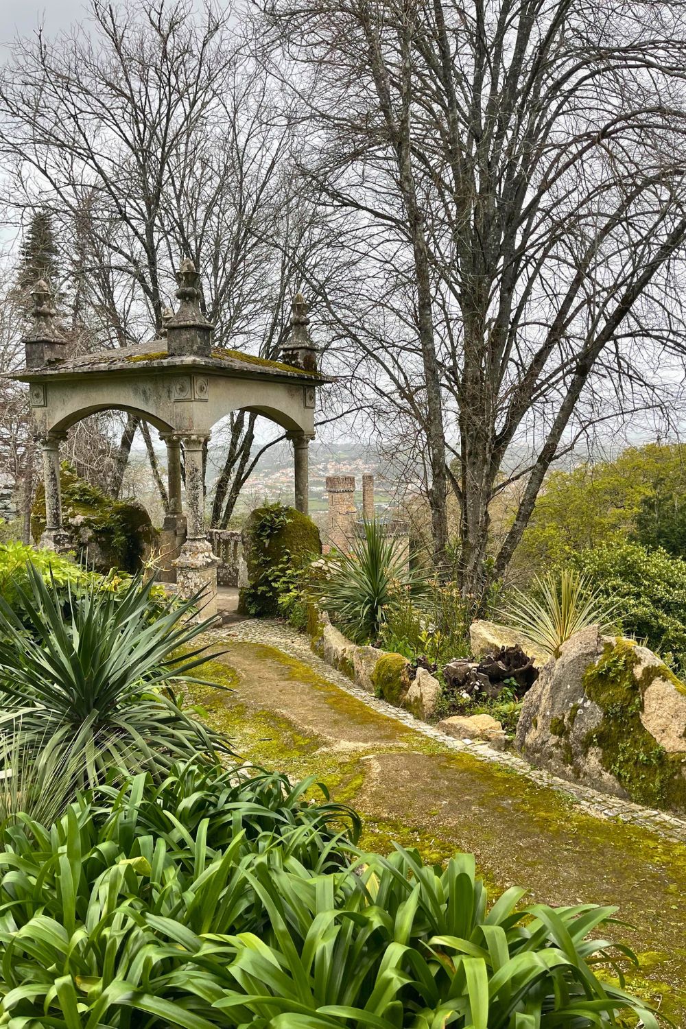 An old stone pergola surrounded by lush greenery and bare trees in what appears to be a tranquil garden setting, with a moss-covered path leading up to it and a view of distant houses in the background, creating a serene and historic atmosphere