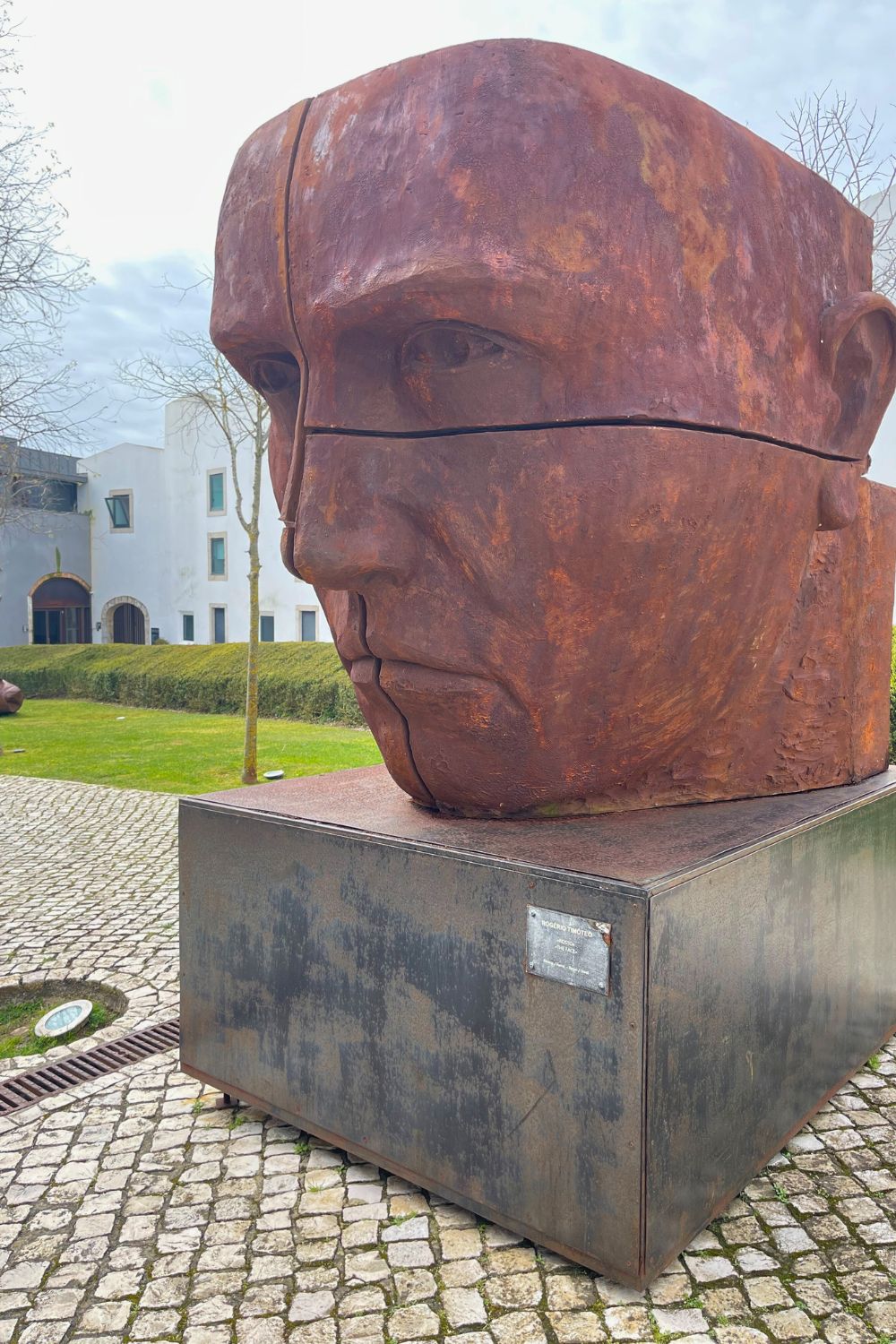 A large, rust-colored sculpture of a human head split into sections, displayed outdoors on a stone-paved area with a sparse grass lawn and part of a modern white building in the background, presenting a striking contrast between art and architecture.
