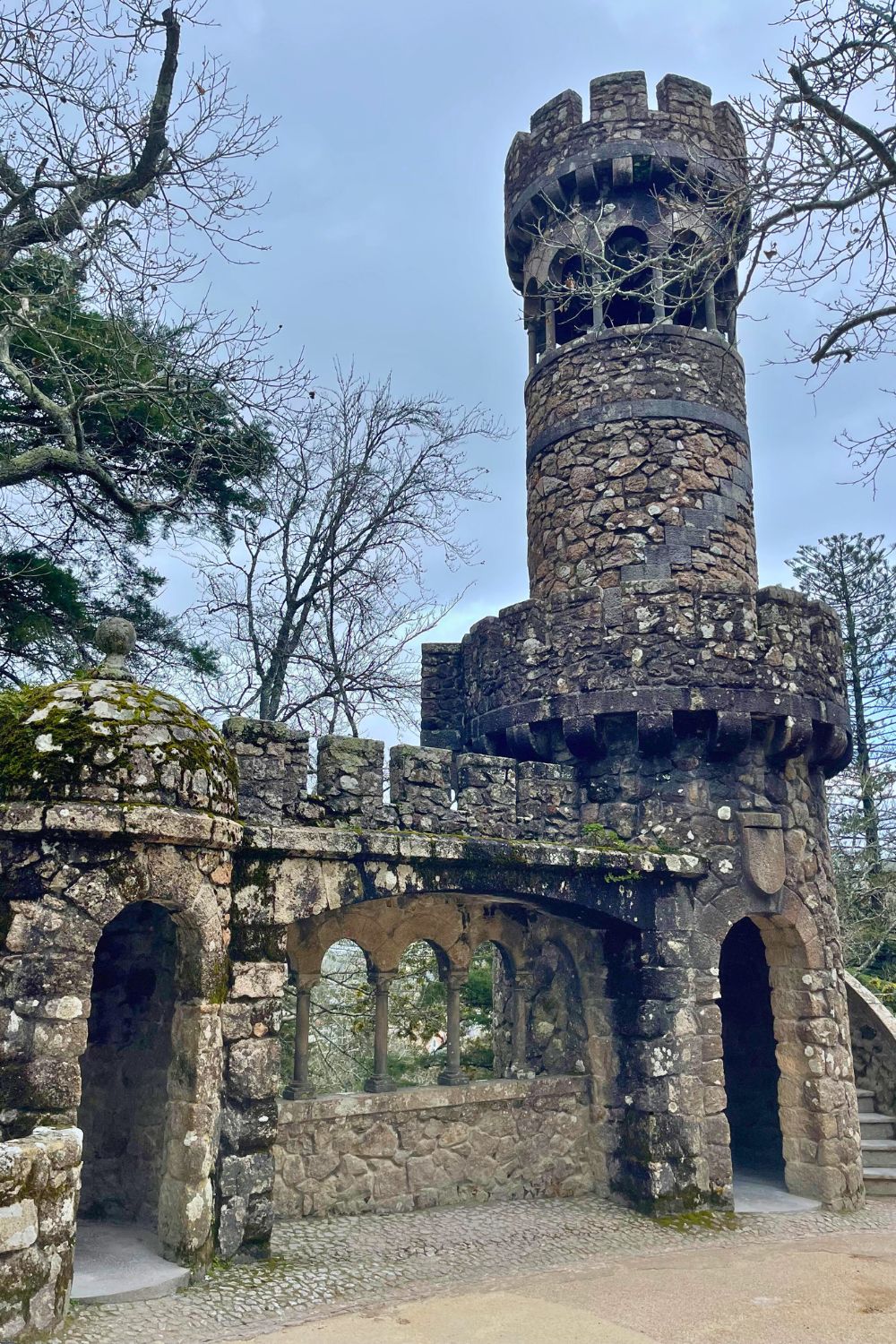 A rustic stone turret with a crenelated top stands guard over a stone archway, evoking a sense of medieval history. The turret is adorned with moss, suggesting age and the merging of nature with architecture, set against a backdrop of bare winter trees.