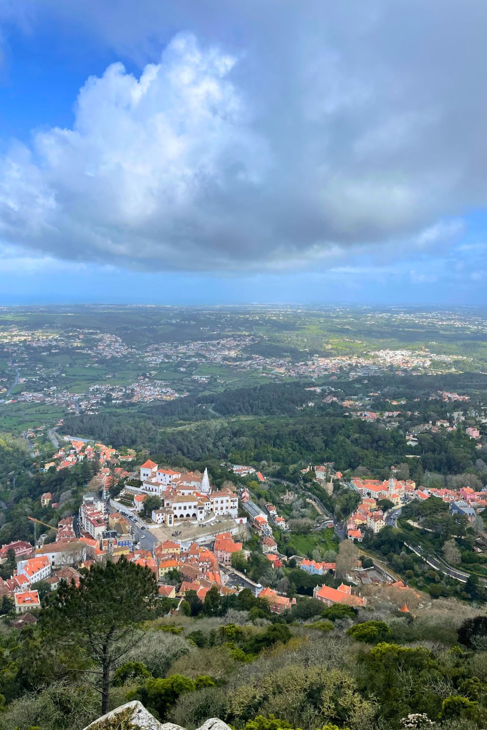 Aerial view of the historic town of Sintra, Portugal, showcasing terracotta-roofed buildings, the prominent National Palace of Sintra with its white facades and twin chimneys, surrounded by lush greenery.