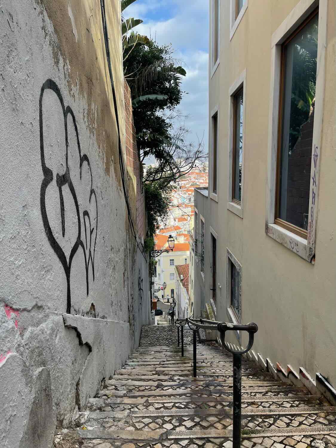 A narrow and steep stairway in Lisbon's old town, flanked by old walls with graffiti, leading down to a picturesque, quiet street.