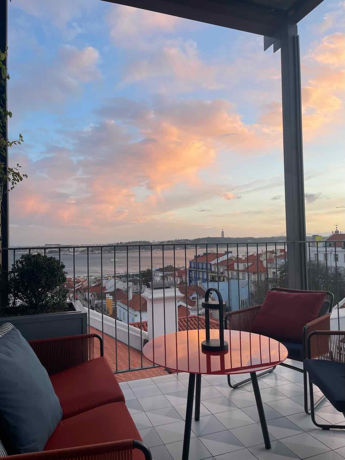 A tranquil rooftop setting in Lisbon at sunset with plush red seating, overlooking a picturesque view of the city under a pastel sky.