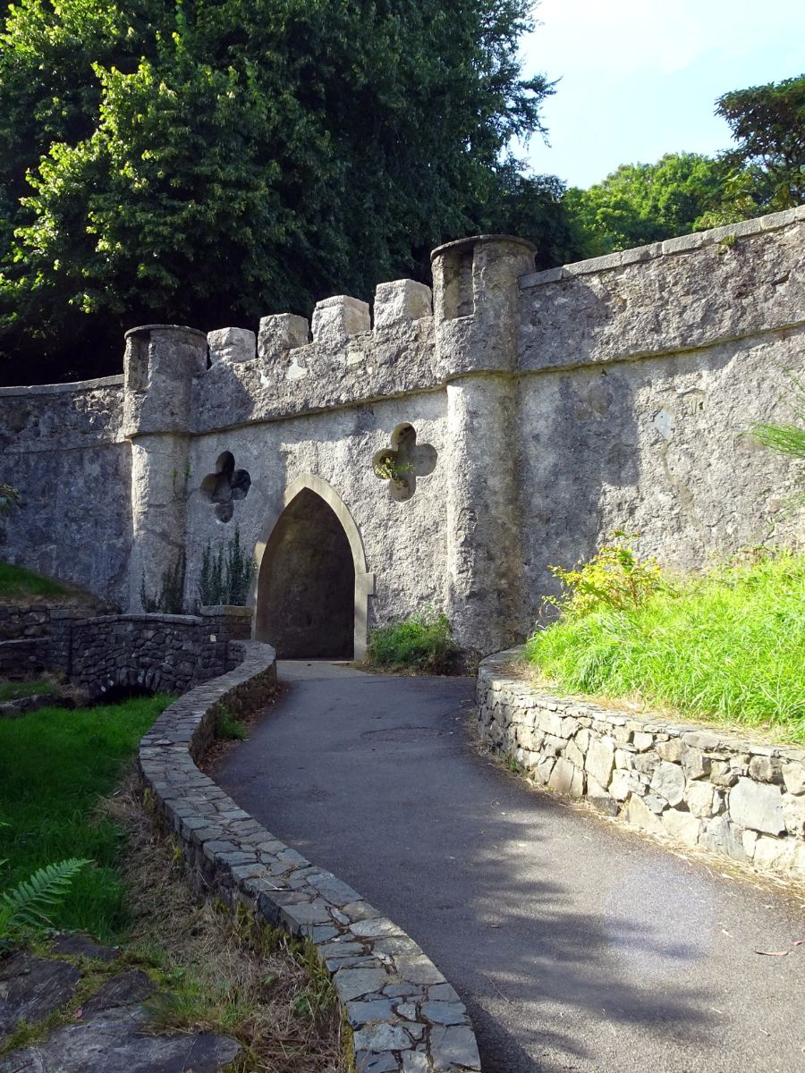 An old stone archway with a crenelated wall on a pathway, suggesting an entrance to an ancient castle or garden. The path curves gently towards the arch, flanked by greenery and shaded by mature trees.