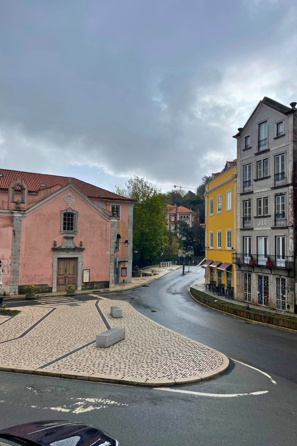 A quiet street in Sintra curves between colorful buildings, including a pastel pink structure with baroque architectural details and a bright yellow building with a cafe on the ground floor. The cobblestone road glistens slightly from recent rain, enhancing the quaint charm of this historic town