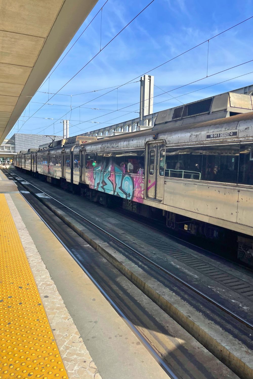 A commuter train adorned with vibrant graffiti stands idle on the tracks at a station, framed by the station's modern, clean architecture and the overhead lines that power its journey. The image captures a moment of stillness in the everyday hustle of city transit.