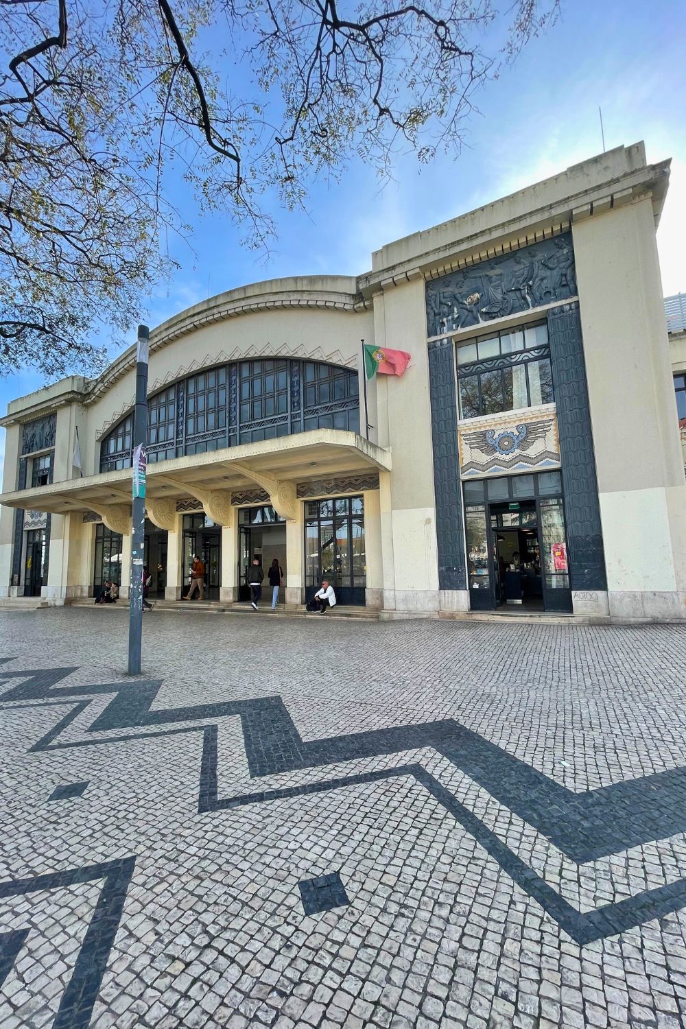 The façade of the Cascais train station presents a beautiful blend of traditional Portuguese architecture and modern functionality. The building, adorned with ornate azulejo tiles and an elegant arched entrance, sits under a soft sky, while the Portuguese flag flutters proudly above. The foreground features the distinctive calçada Portuguesa, a traditional-style cobblestone pavement creating a captivating geometric pattern.