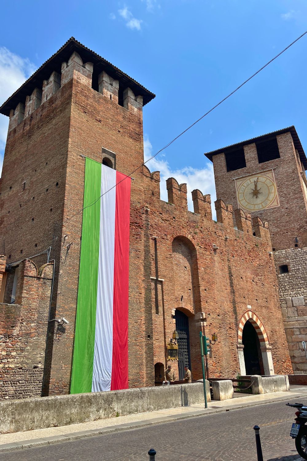 A historic brick building in Verona with a large Italian flag hanging vertically from the top. The building features crenellated battlements and a tower with a large clock face, set against a clear blue sky.