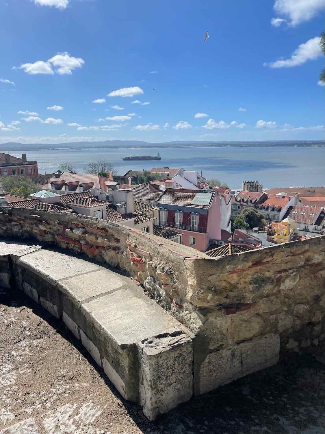 A scenic view of Lisbon's traditional architecture and the Tagus River in the background, captured from a high viewpoint on a sunny day.
