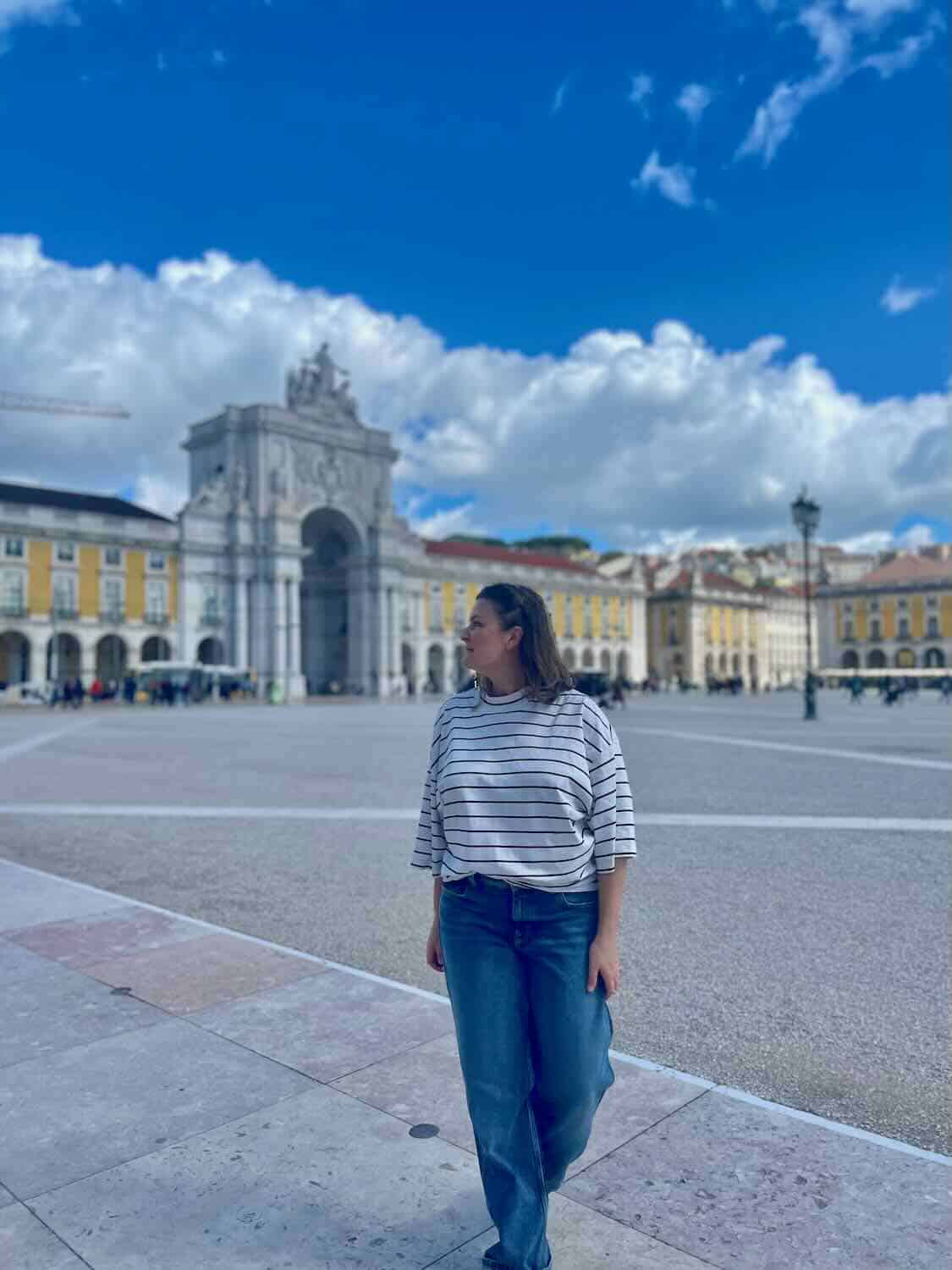 A woman strolling in front of Lisbon's expansive Praça do Comércio with the notable Rua Augusta Arch, under a sky with fluffy clouds