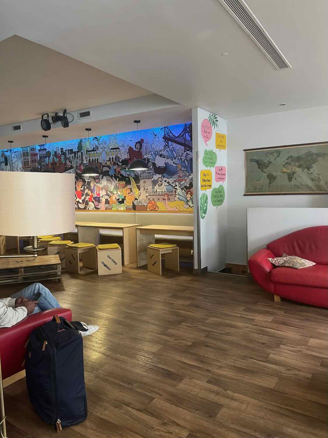 A cozy common area inside a Lisbon hostel, with a traveler resting on a red sofa, and a vibrant wall mural showcasing the city's landmarks