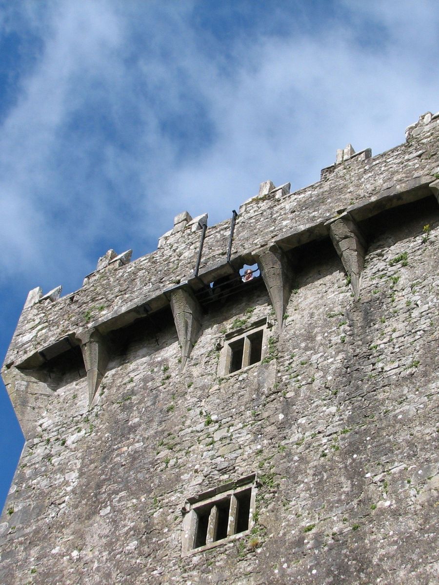 A view from below of a stone castle tower against a clear blue sky, with a person leaning out of a niche, appearing small in comparison to the structure's massive walls.