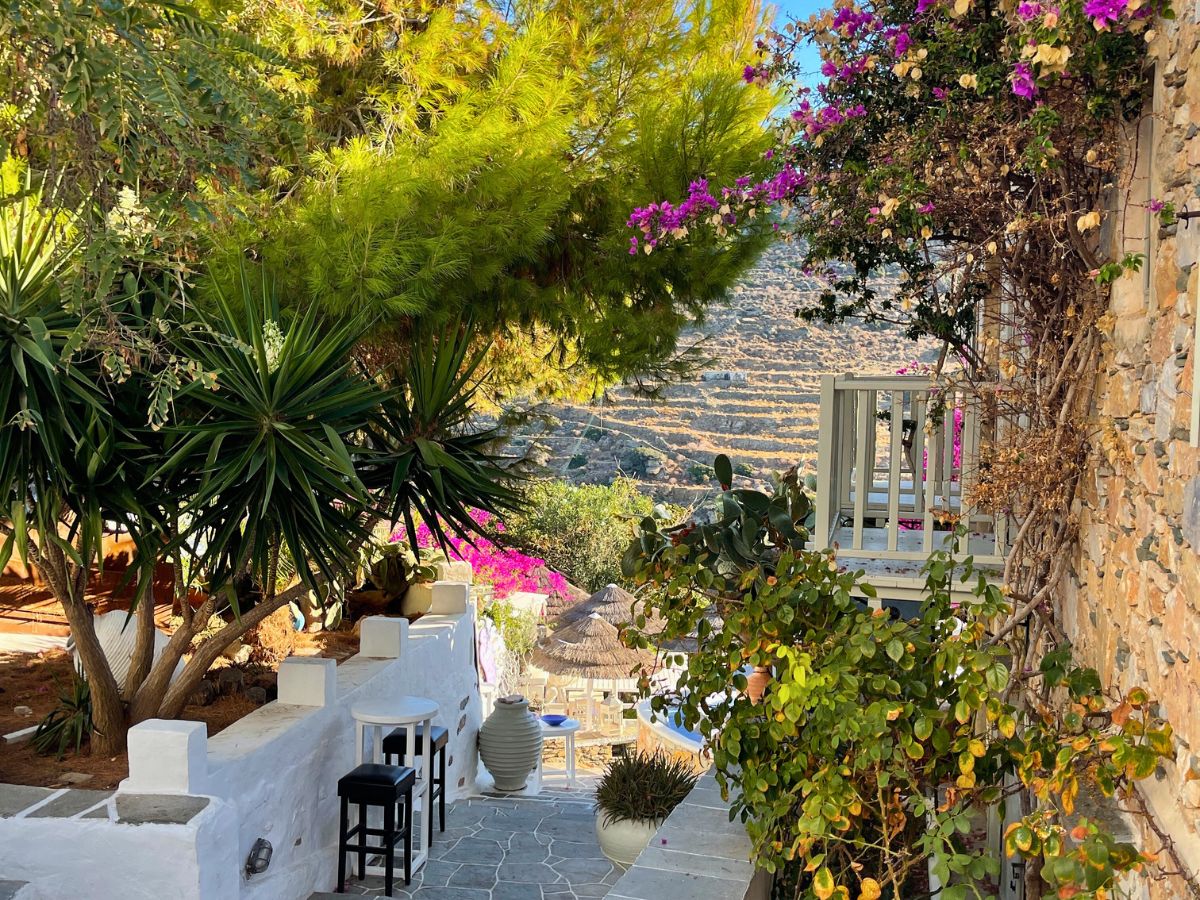 A picturesque garden pathway in Sifnos, lined with lush greenery, flowering plants, and a white-washed building, offering a glimpse into the tranquil beauty of the island's accommodations.