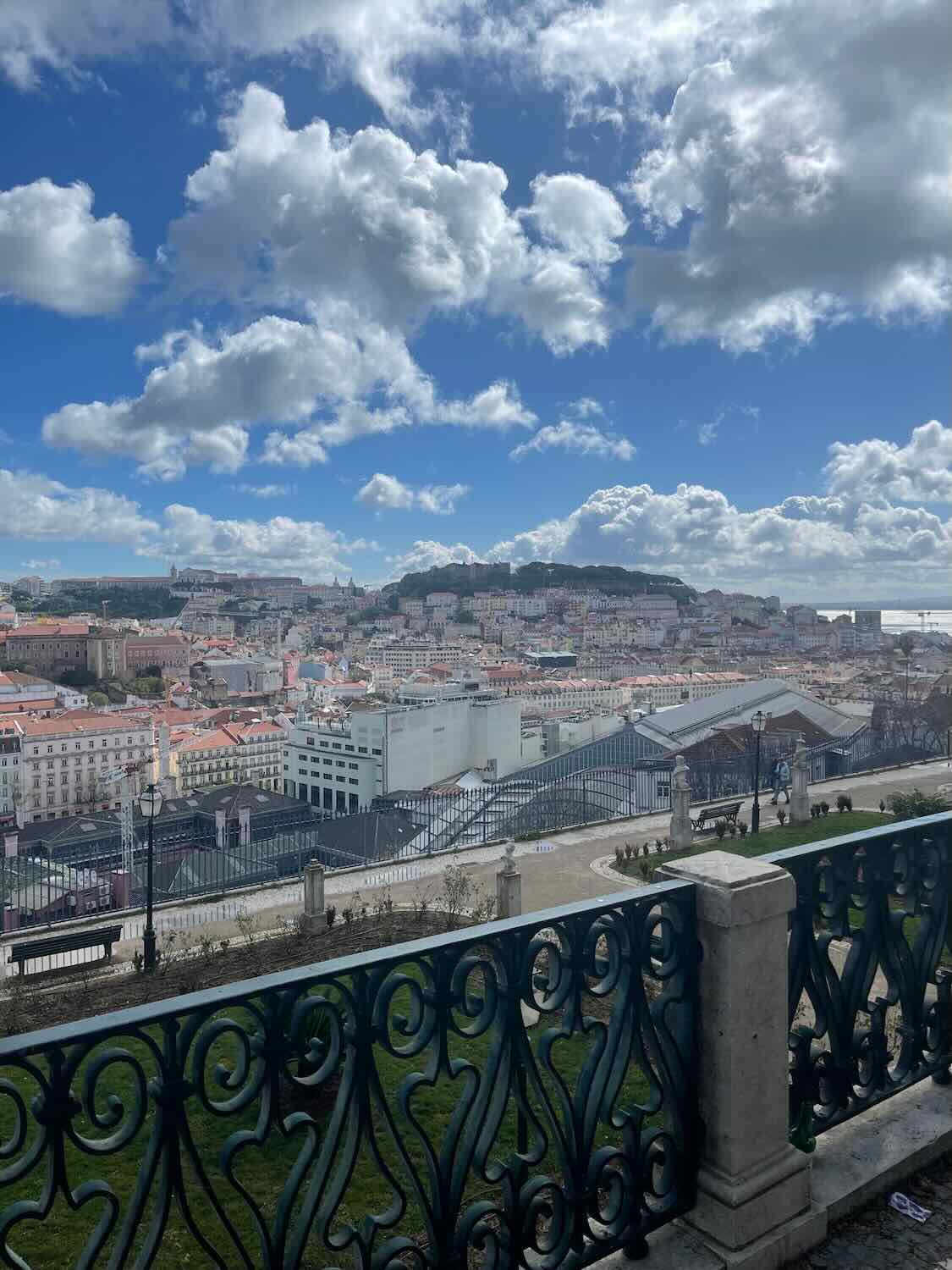 A panoramic view from a vantage point in Lisbon, showing the cityscape with its historic buildings under a dramatic cloud-filled sky.