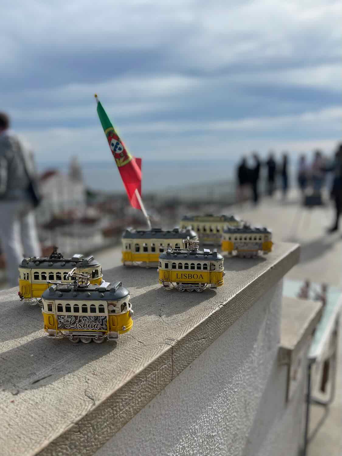 Miniature yellow trams with 'Lisboa' branding displayed on a ledge against the backdrop of a blurred Portuguese flag, symbolizing Lisbon's iconic transport