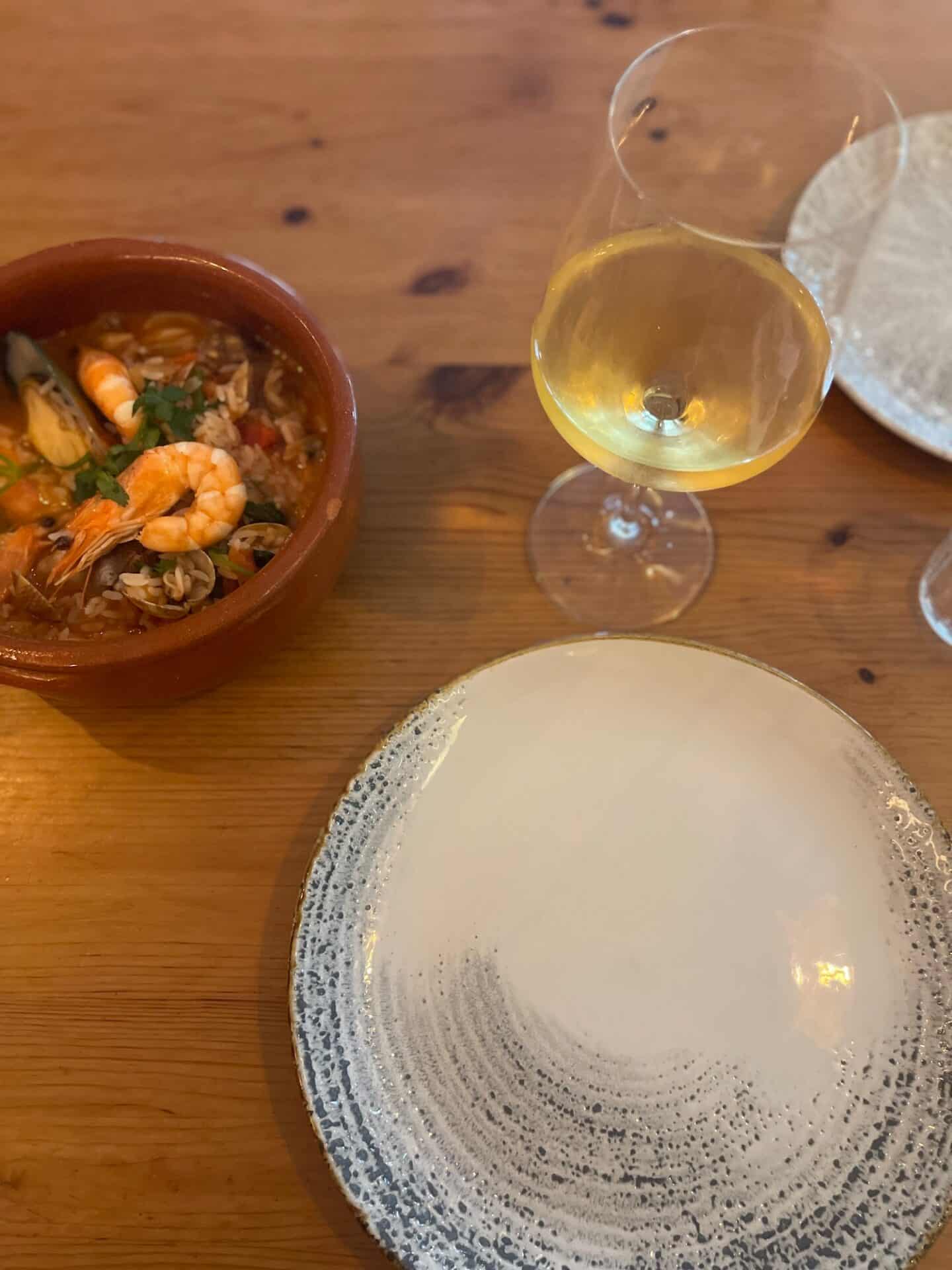 A traditional Portuguese seafood dish served in a terracotta bowl next to a glass of white wine, with an empty decorative plate, on a wooden table, capturing the flavors of Tavira's culinary offerings
