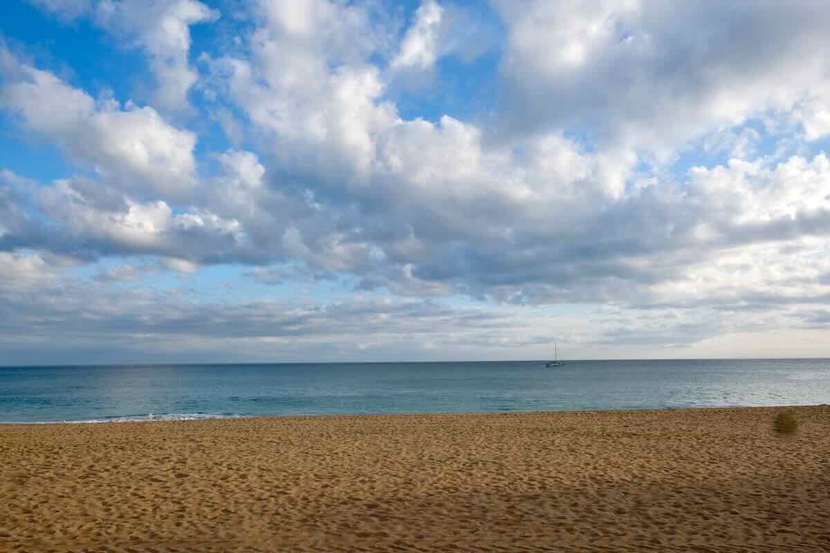 A serene beach scene with golden sands in the foreground, calm blue ocean waters stretching to the horizon, and a sailboat in the distance under a sky dotted with fluffy clouds.