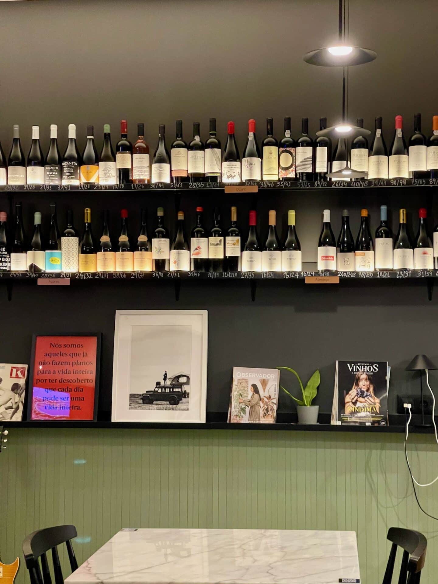 A well-lit wine bar interior featuring a variety of wine bottles on shelves, framed artwork, and magazines on a wall-mounted ledge above a marble table, reflecting the cozy and cultured ambiance of Tavira's local dining scene.