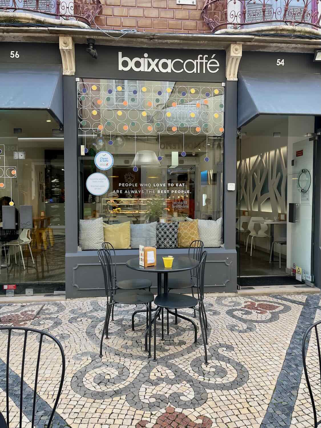 Entrance to Baixa Café in Faro, with its modern gray storefront and decorative window design featuring circles of multiple colors; outdoor seating available on a mosaic-tiled sidewalk, a sign quoting 'People who love to eat are always the best people.