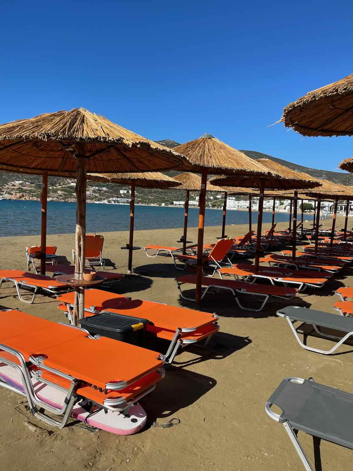 Orange sunbeds and natural straw umbrellas line the sandy shore of Sifnos, inviting relaxation on a sunny day, a serene addition to any Sifnos itinerary.