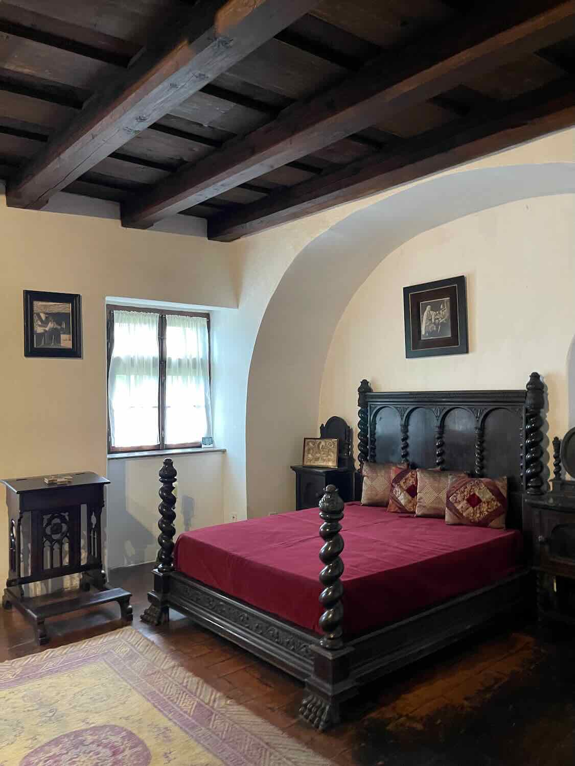 A grand, antique wooden bed with spiral bedposts and a red duvet in a room with exposed wooden ceiling beams, an archway, and traditional furnishings, suggesting a historical setting.