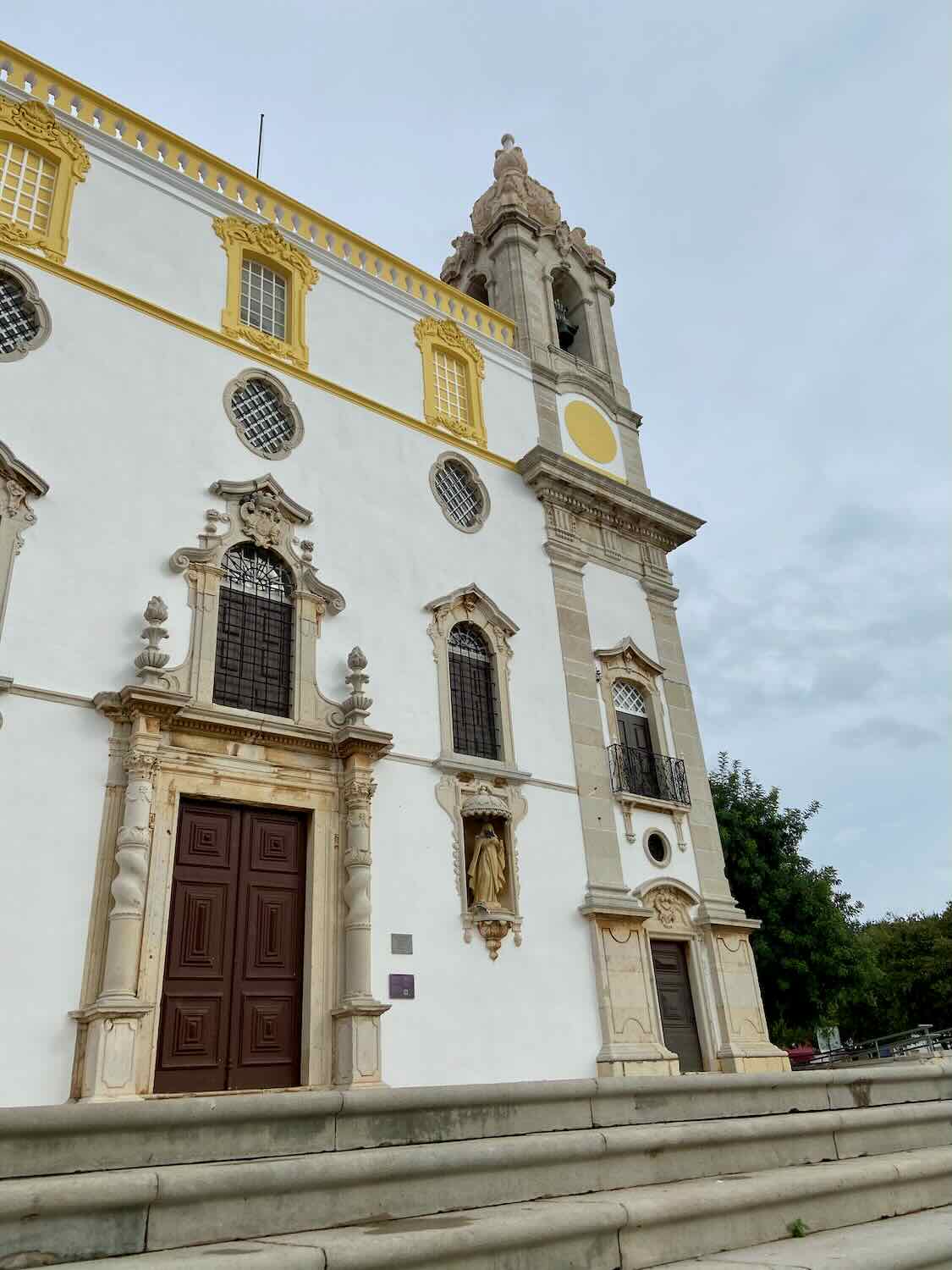The striking façade of Carmo Church in Faro, displaying intricate Baroque architectural details with contrasting yellow trim around windows and ornate stone carvings, set against a cloudy sky.