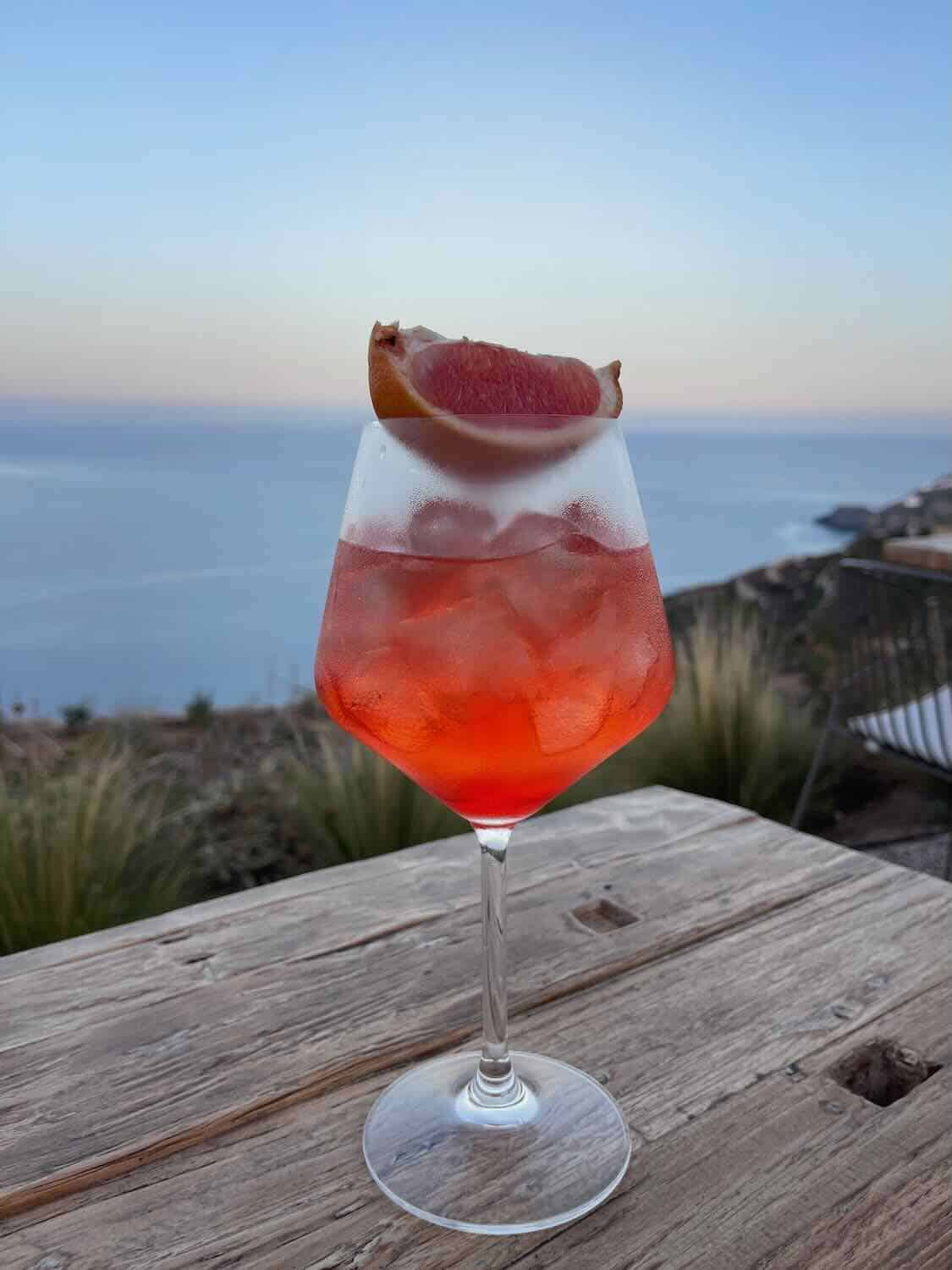 A refreshing cocktail adorned with a slice of grapefruit, resting on a rustic wooden table with a scenic view of the Sifnos coastline, a perfect sip stop on a Sifnos itinerary.