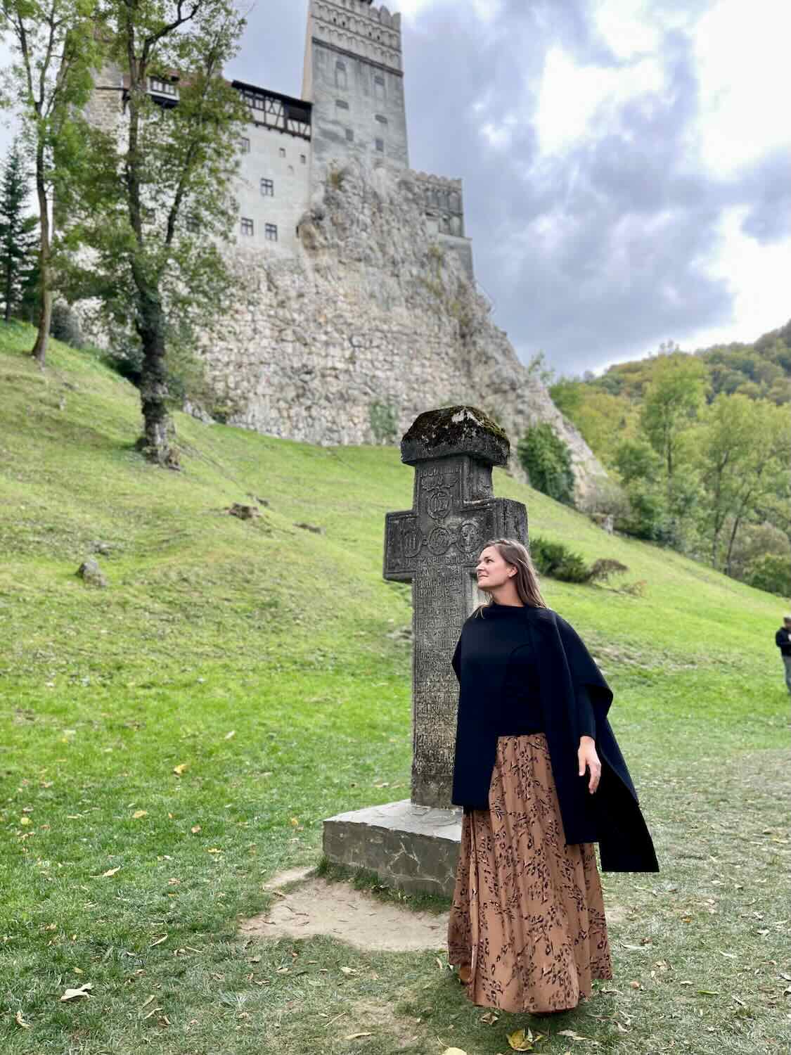 A woman in a black cape and patterned skirt stands contemplatively by a stone cross, with the towering, grey walls of Bran Castle rising in the background amidst lush greenery and a moody sky.