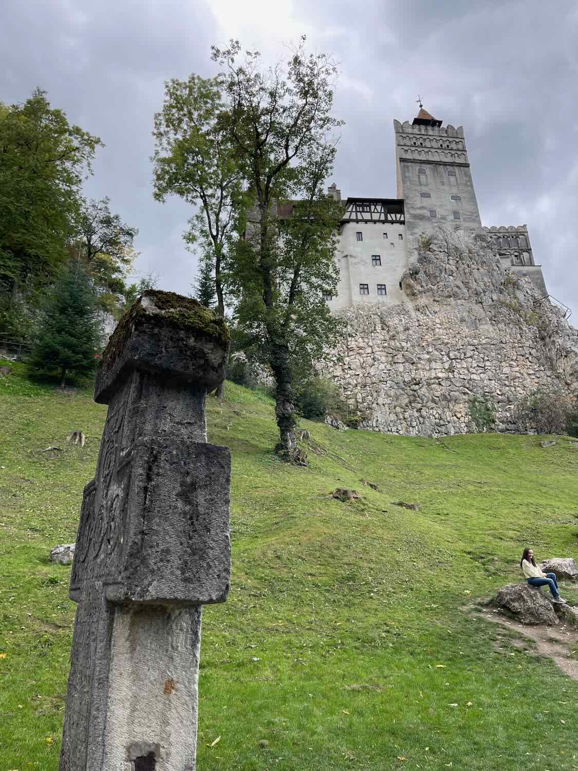 Imposing view of Bran Castle perched on a rocky outcrop against an overcast sky, with a stone cross monument in the foreground and trees surrounding the scene, capturing the castle's majestic and mysterious ambiance