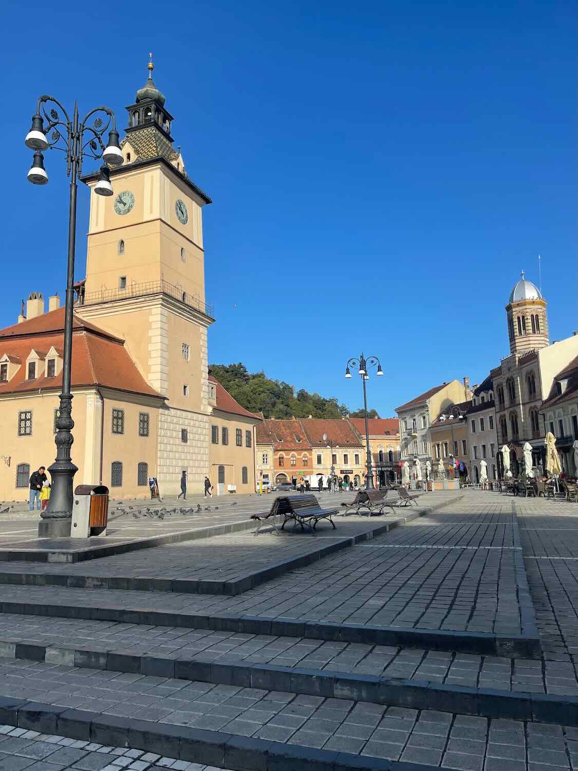 Bright, sunny day over Brasov's town square, showcasing the old town's charming architecture with the Black Church's tower and ornate street lamps, all against a vivid blue sky.