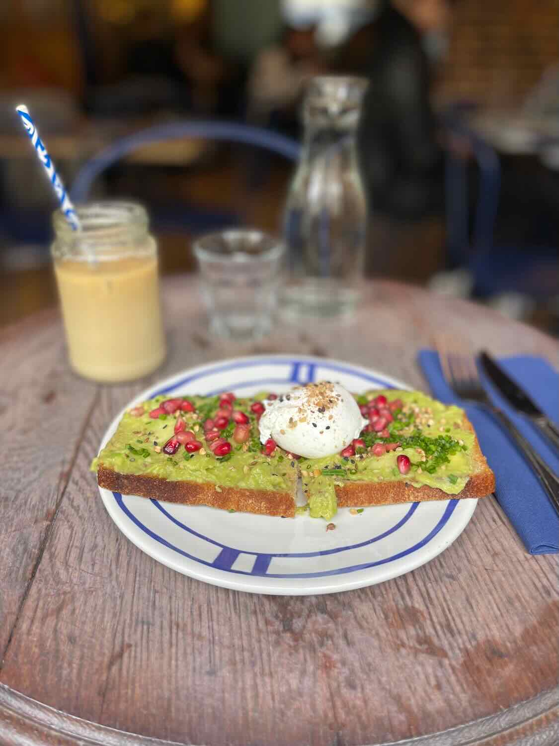 A beautifully presented avocado toast topped with pomegranate seeds and a poached egg, served on a striped blue and white plate, accompanied by a refreshing iced latte on a rustic wooden table.