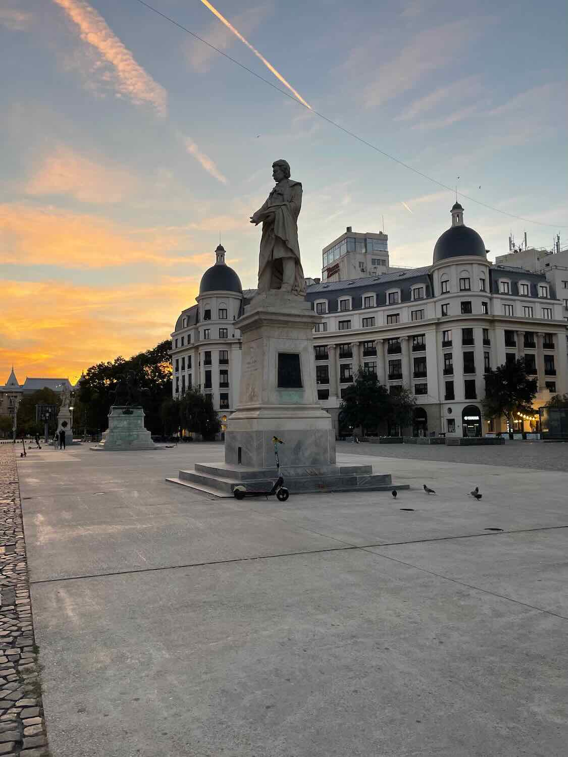 Dusk sets over Bucharest as the silhouette of a statue dominates the foreground and pastel-colored skies backdrop the city's architecture, with buildings and street lights beginning to glow.