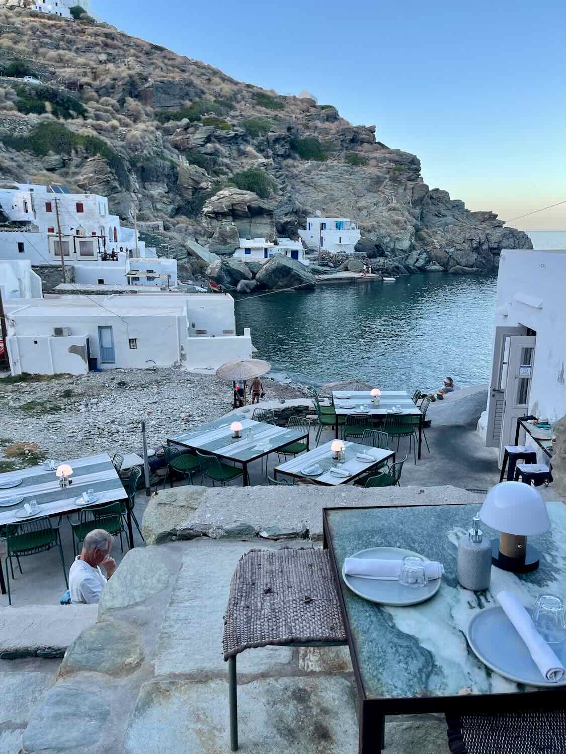 Tables set for a waterfront dining experience in Sifnos, nestled among white-washed buildings with the Mediterranean Sea as a backdrop
