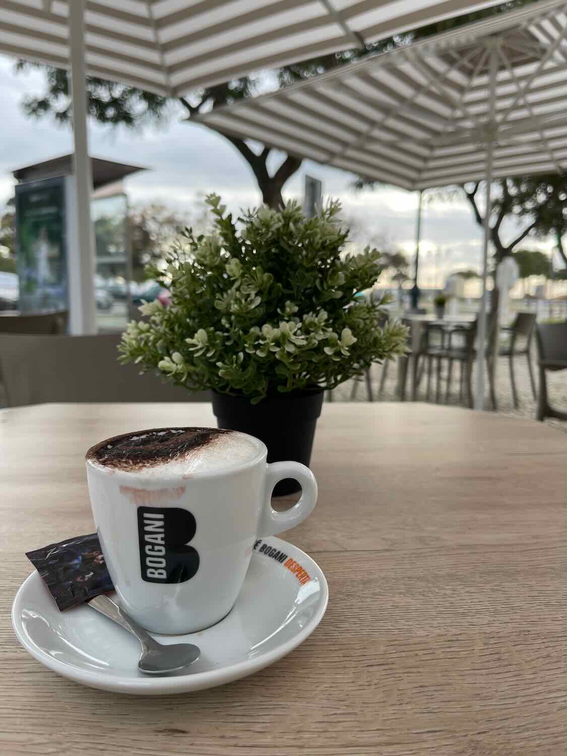 Close-up of a cappuccino in a white cup with the logo 'BOGANI' on a wooden table, accompanied by a sugar packet, with a potted green plant in the background and an outdoor patio setting with white pergola overhead.