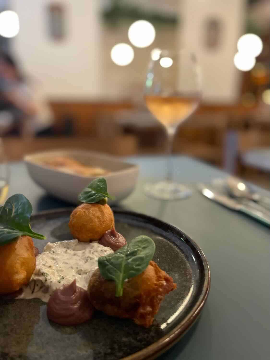 An intimate dining scene featuring deep-fried gourmet bites with dipping sauces on a ceramic plate, focused sharply in the foreground. A glass of rosé wine adds a touch of elegance to the relaxed ambiance of the restaurant, with blurred background details creating a cozy dining atmosphere.