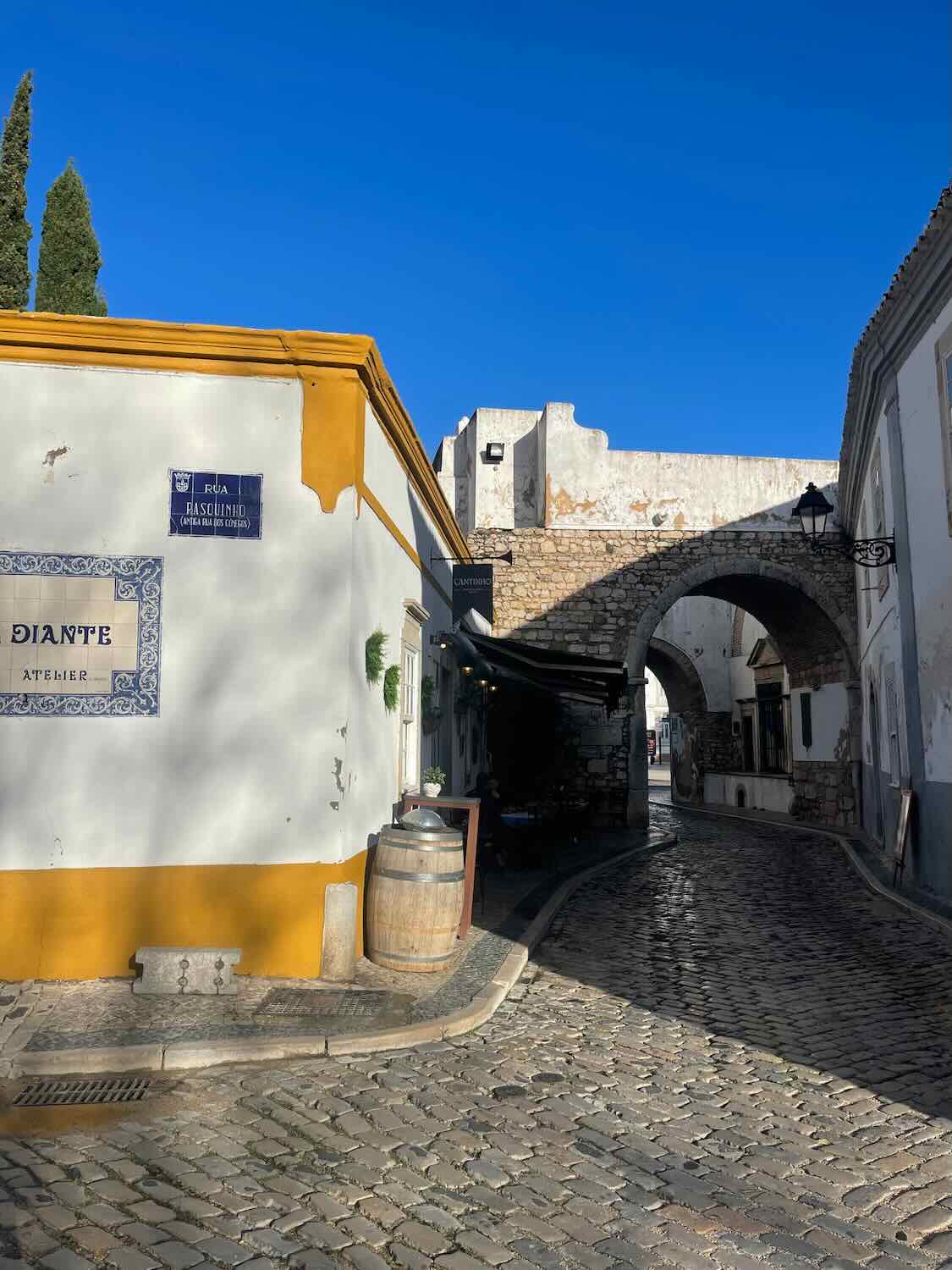 Historical street in Faro featuring the Arco da Vila with its traditional architecture, a blue street sign for 'Rua Rasquinho,' and a quaint corner adorned with a ceramic plate and a wooden barrel.